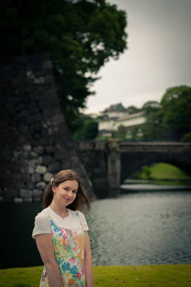 Young woman with long brown hair wearing a white t-shirt and a colorful floral dress, standing near a body of water with a stone bridge and traditional Japanese castle in the background.