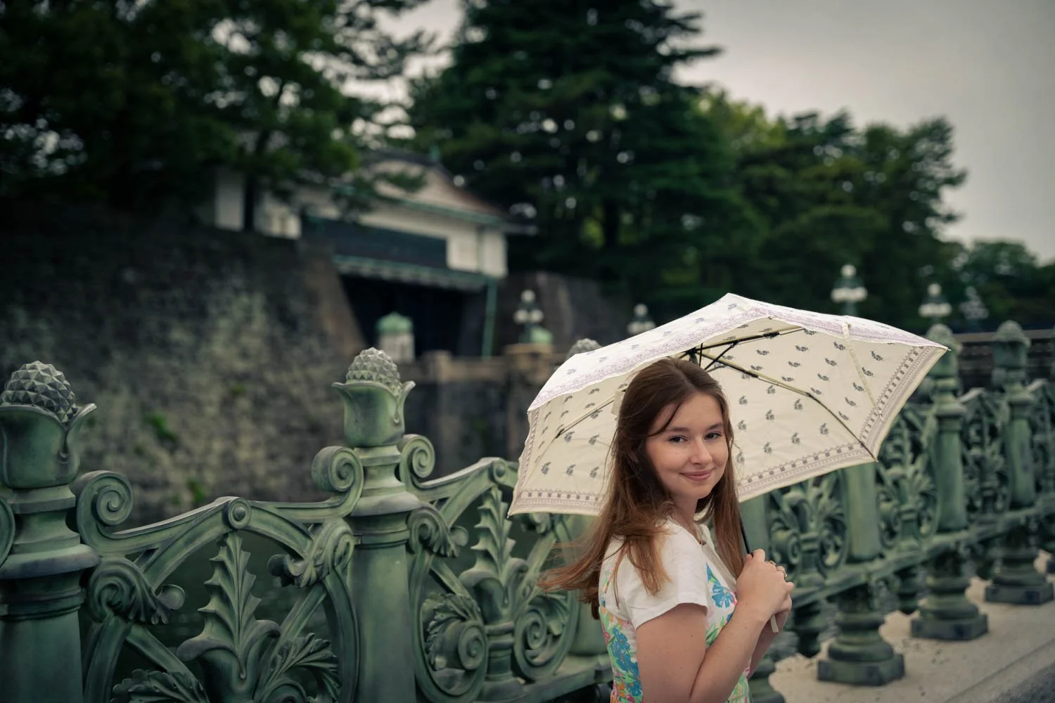 Young woman holding a patterned umbrella, smiling, standing next to an ornate green metal fence with a historic building and lush trees in the background on a cloudy day.