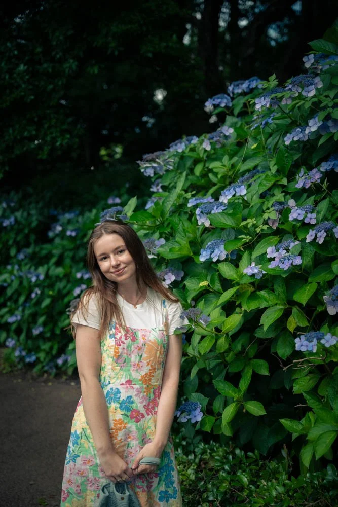 A young girl wearing a floral dress standing next to a lush bush with purple flowers in a garden or park.