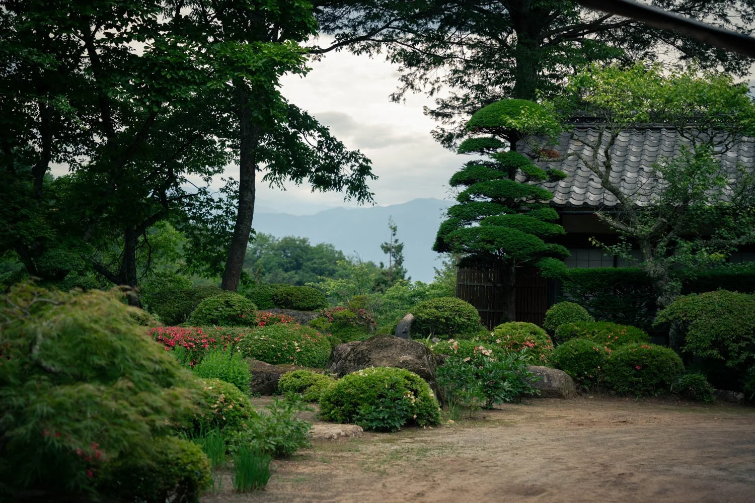 Traditional Japanese garden with neatly trimmed bushes, rocks, colorful flowers, and a wooden building with a tiled roof surrounded by trees, with mountains in the background