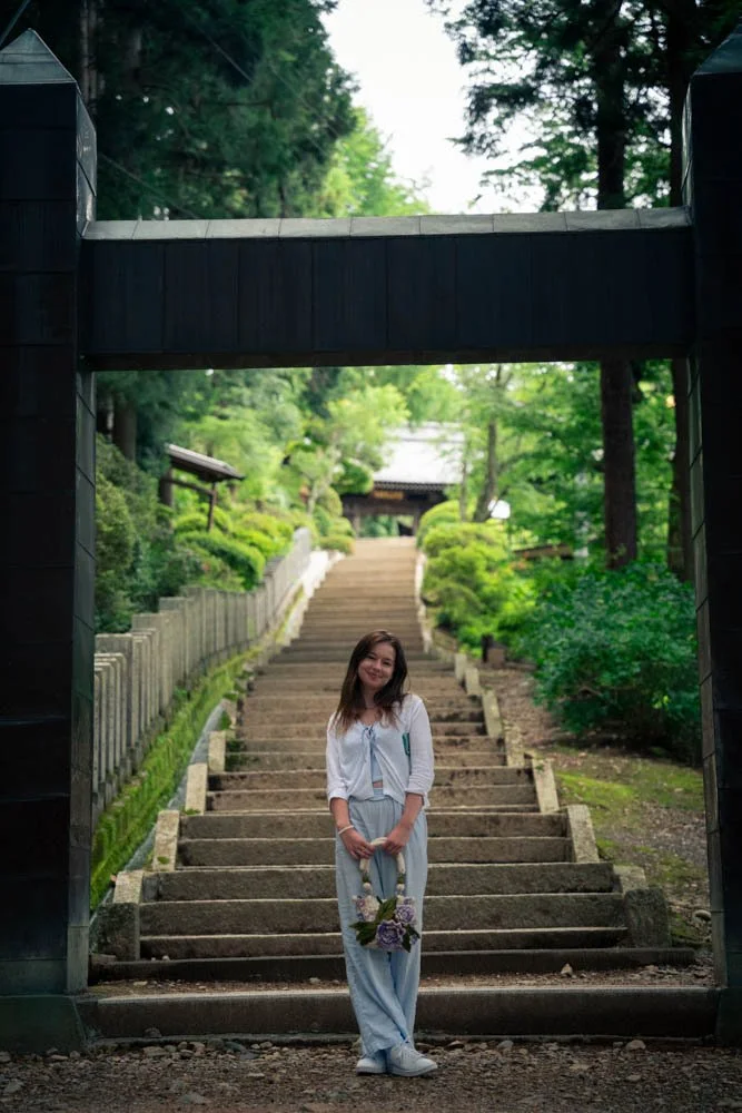 A young woman standing on stone steps in a lush green garden, holding a bouquet of flowers, smiling at the camera.