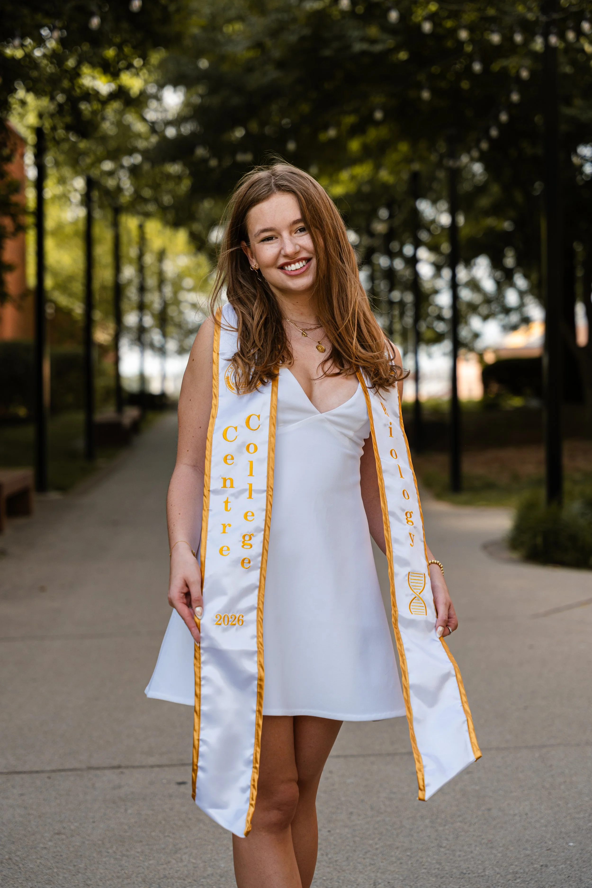 Young woman smiling at camera wearing a white graduation gown and yellow stole with '2026' embroidered, outdoors in park with trees in background.