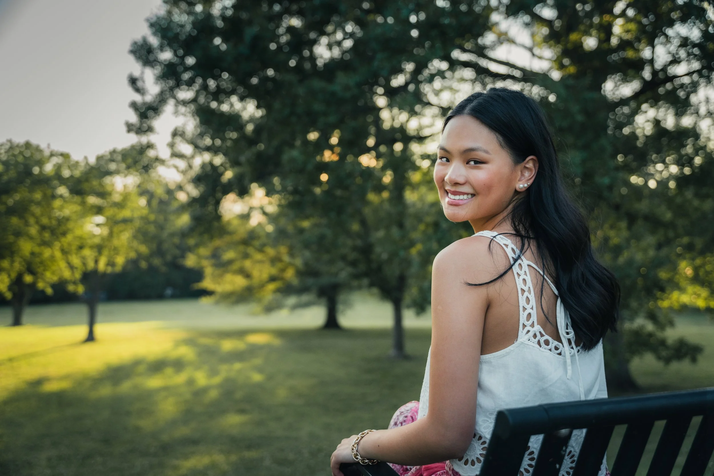 A young woman with long dark hair smiling while sitting on a park bench with trees in the background during late afternoon or early evening.