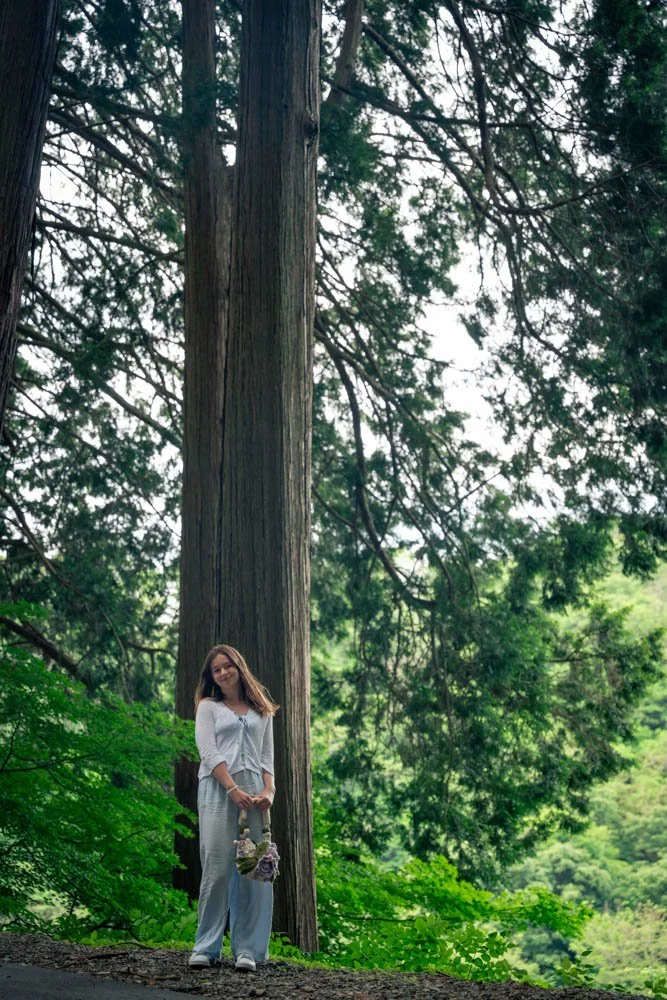 A woman standing outdoors near a large tree, holding a small bouquet of flowers, surrounded by green foliage.