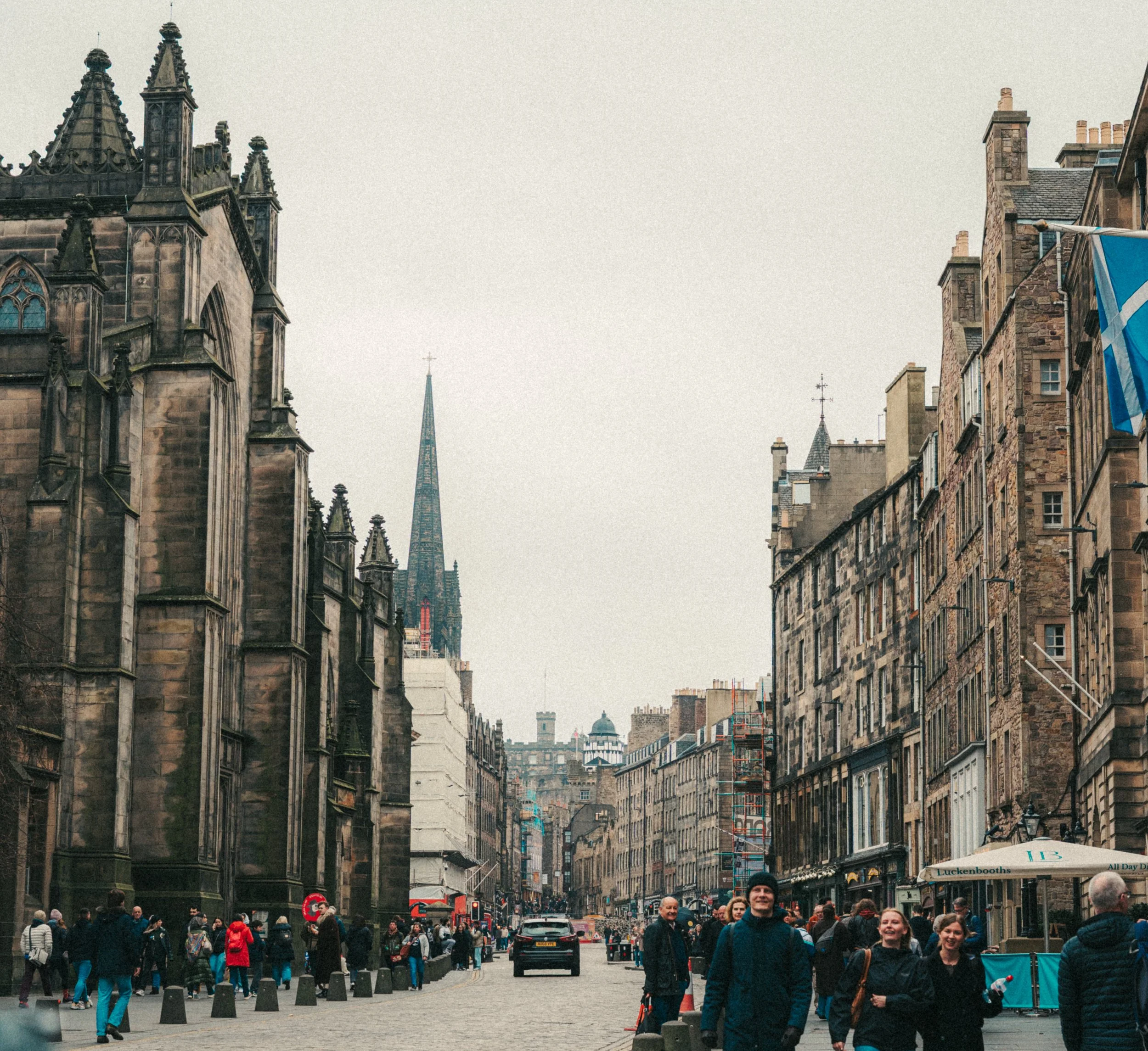 Crowd of people walking on a busy city street with historic buildings and scaffolding, with the spire of a church or cathedral in the background.