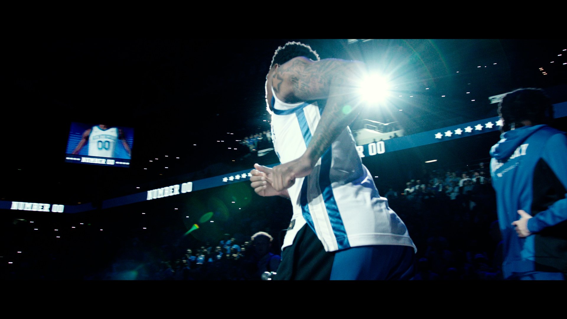 A basketball player in a white and blue jersey on the court, with one hand clasped with a teammate, illuminated by bright stadium lights.