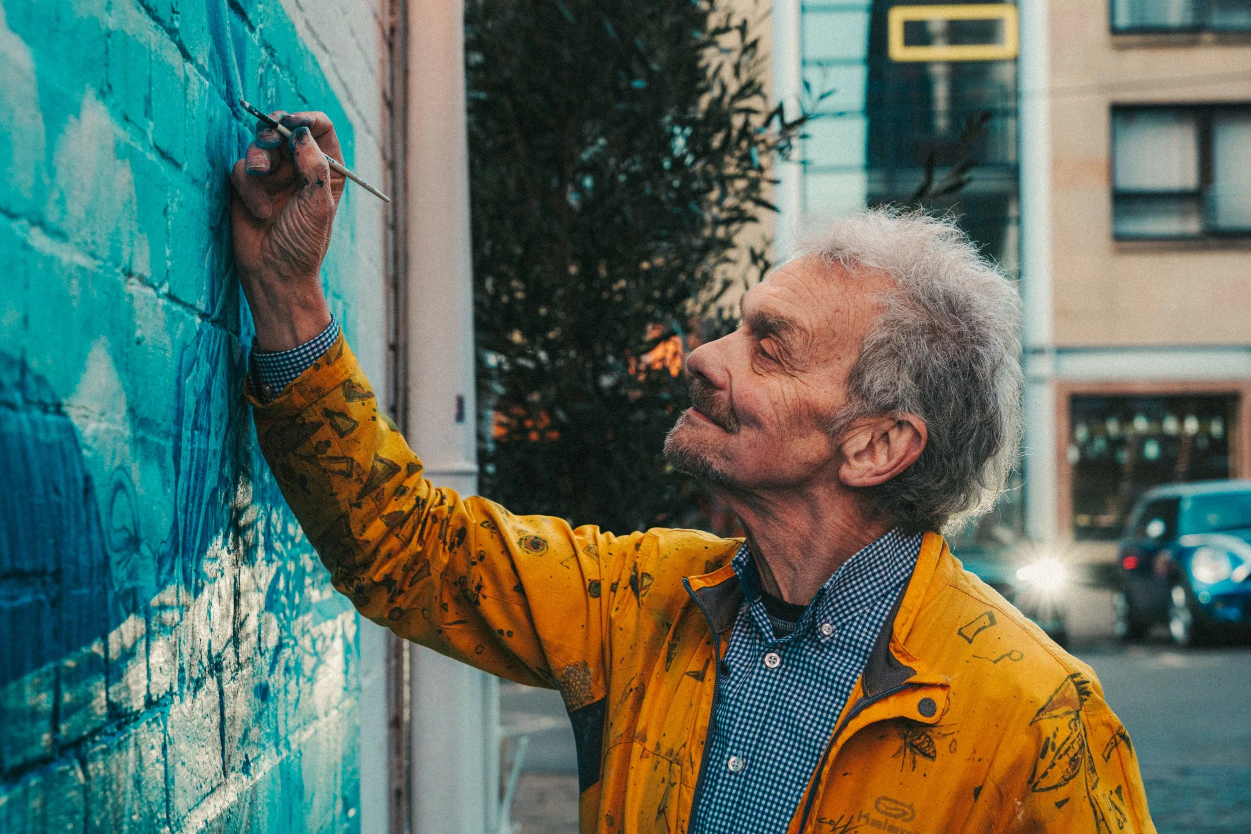 An elderly man with grey hair and a beard painting a mural on a brick wall outdoors. He is wearing a yellow jacket with paint splatters and a blue checkered shirt underneath, smiling softly as he works.