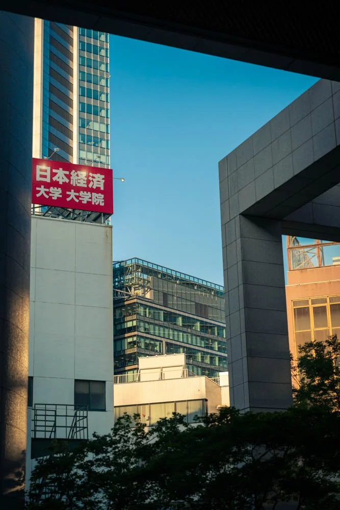 Urban cityscape with modern glass and concrete buildings under a clear blue sky, with a red sign containing Japanese characters on one building.