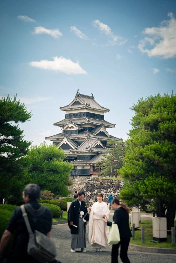 A traditional Japanese multi-tiered castle with a curved roof, surrounded by lush green trees, under a partly cloudy sky. Several people, including two women in kimonos, are walking and talking in the foreground.