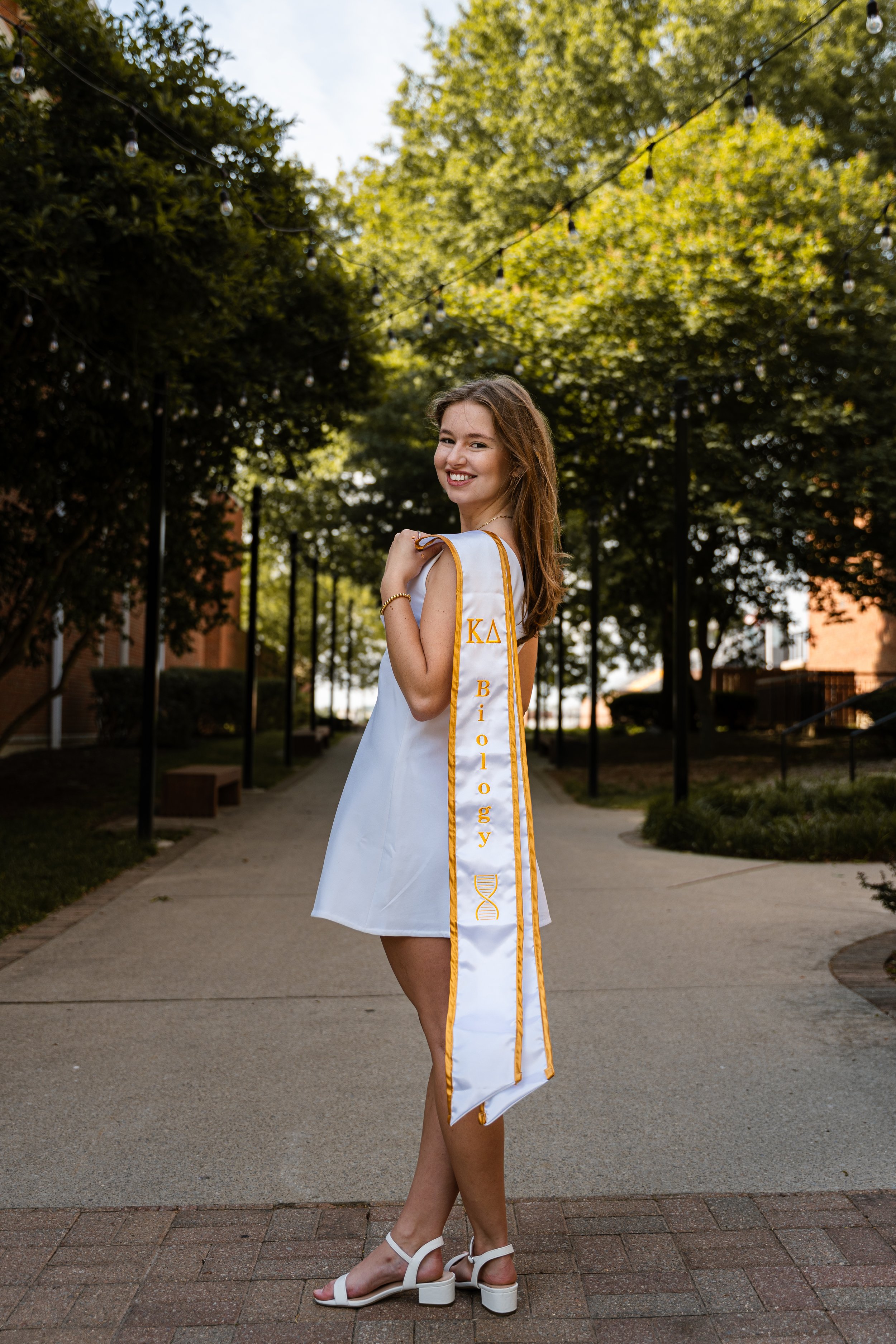 A young woman wearing a white graduation gown and sash with gold trim and Greek letters standing on a pathway in a park-like setting. She is smiling and looking over her shoulder, with trees and string lights overhead in the background.