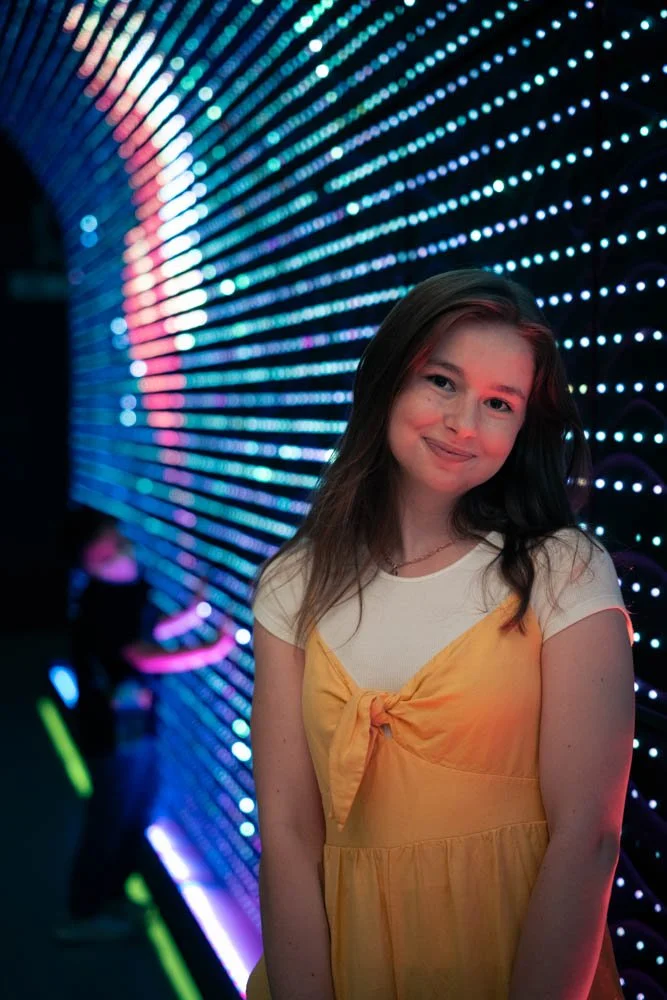 A young woman with long brown hair wearing a yellow dress with a tied front over a white shirt, standing in front of a vibrant, colorful LED light wall at an indoor location, smiling at the camera.
