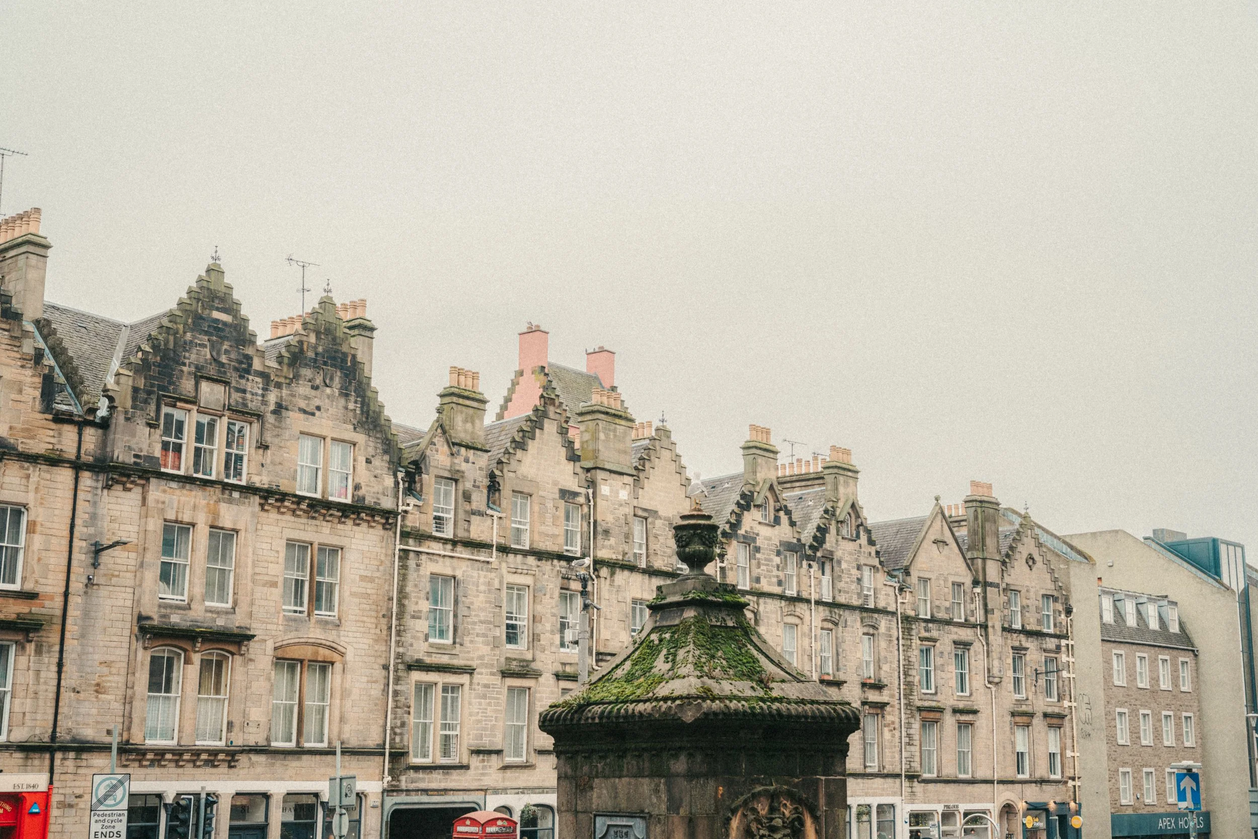 Row of historic stone buildings with tall, narrow windows and decorative rooftops, in an urban setting, with a fountain in the foreground and signs for traffic and pedestrians.