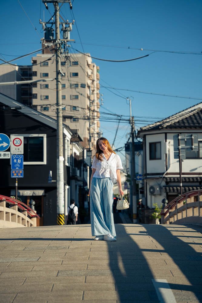 A woman in casual clothing standing on a small bridge over a street in a city neighborhood, holding a handbag, with buildings and power lines in the background under a clear blue sky.