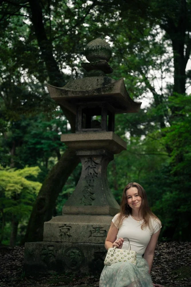 A young woman with long brown hair, wearing a white top and a light colored skirt, is sitting and smiling at the camera in a wooded area.