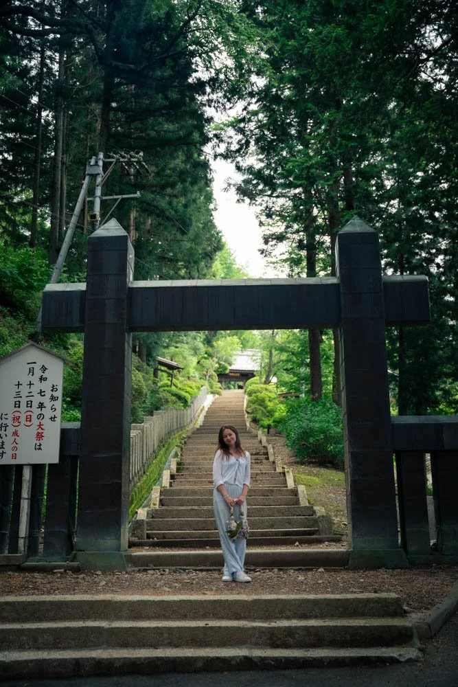 A woman standing at the bottom of a wide stone staircase leading up through a lush green forest, with a black traditional Japanese torii gate at the entrance, holding a bouquet of flowers.