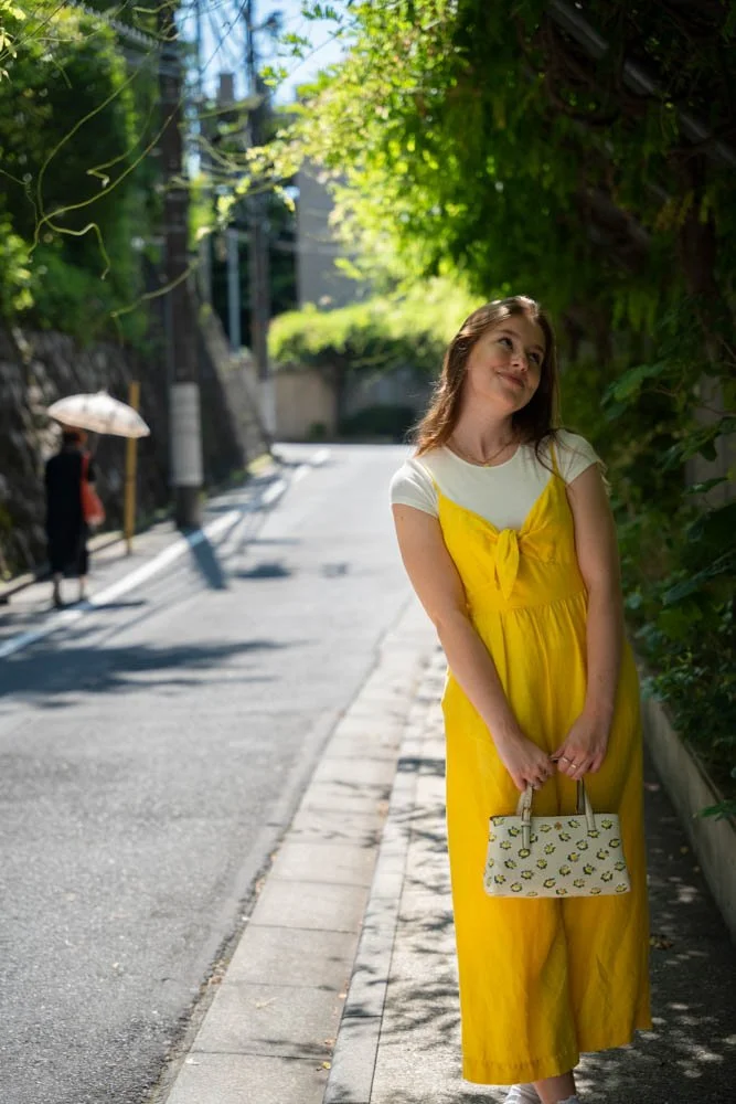 A woman in a yellow dress standing on a sunny sidewalk, holding a white bag with yellow flower patterns, with greenery and a person holding an umbrella in the background.
