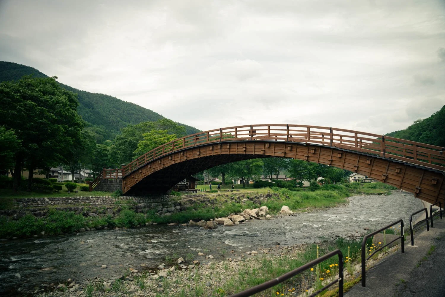 A wooden arched footbridge over a rocky river in a park with green trees and distant hills under a cloudy sky.