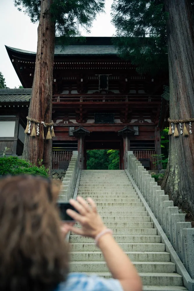 Person taking a photo of a traditional wooden Japanese shrine with stairs and large trees surrounding it.