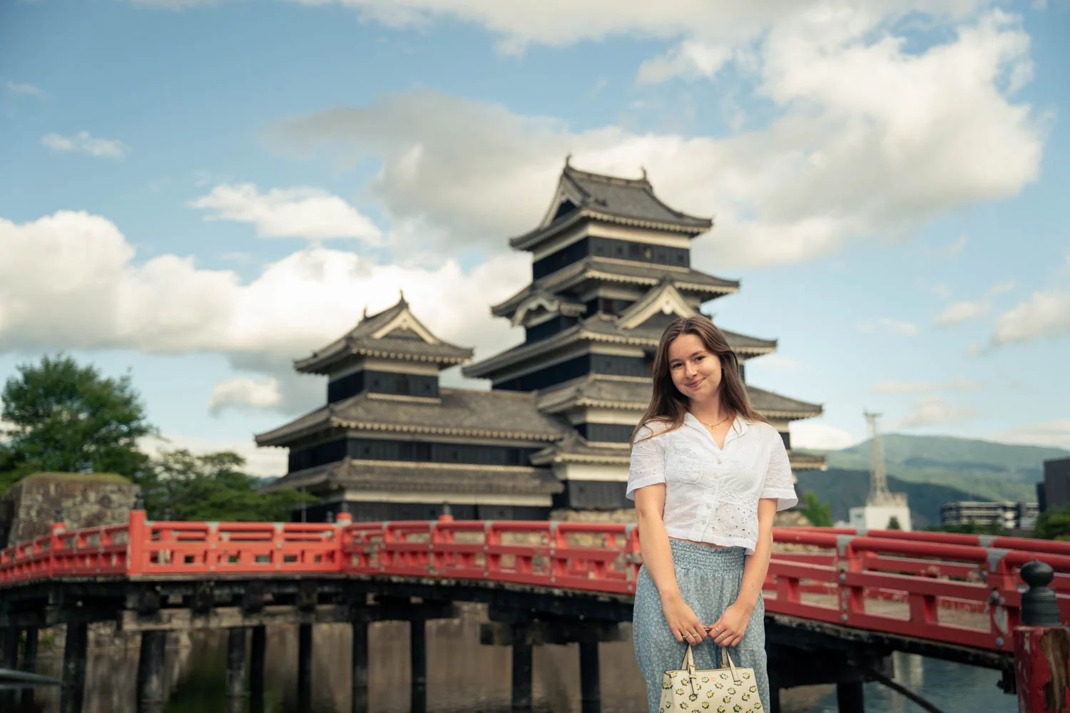 Young woman in white blouse and blue skirt holding a patterned bag standing on a red bridge with a traditional Japanese pagoda-style building and a cloudy sky in the background.
