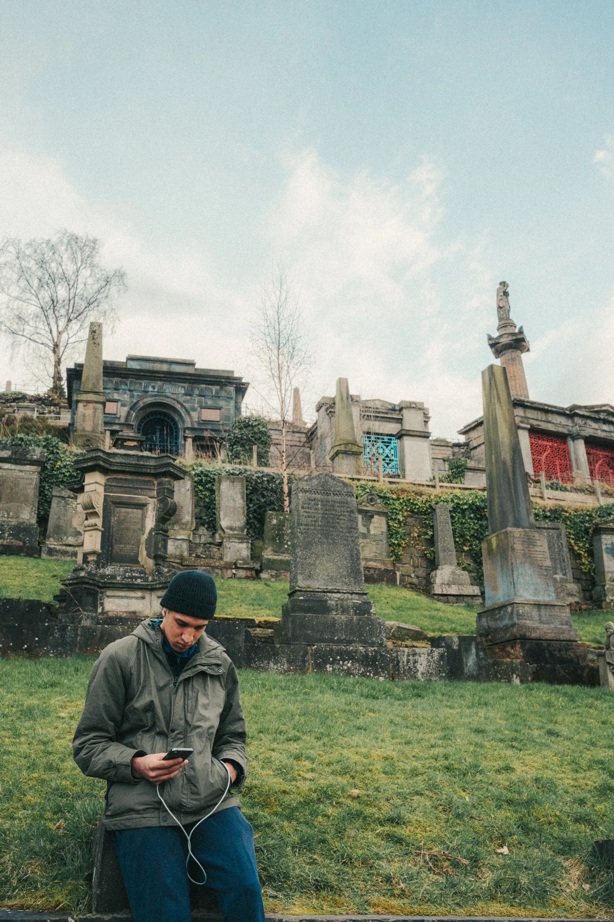 A young man in a green jacket and black beanie is sitting outdoors on green grass, looking at his phone with earbuds connected. Behind him is a hillside with old, weathered tombstones and mausoleums, and a tall statue with a column and a figure at th