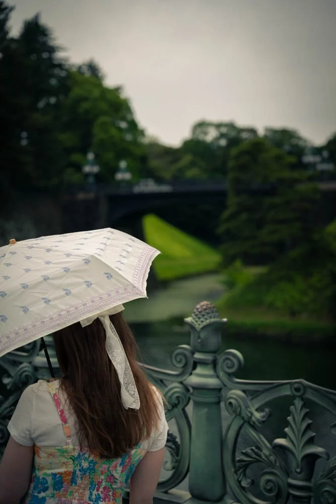 A girl with long hair holding a white umbrella with purple floral patterns, facing away, standing on a decorative bridge over a river with greenery and trees in the background.