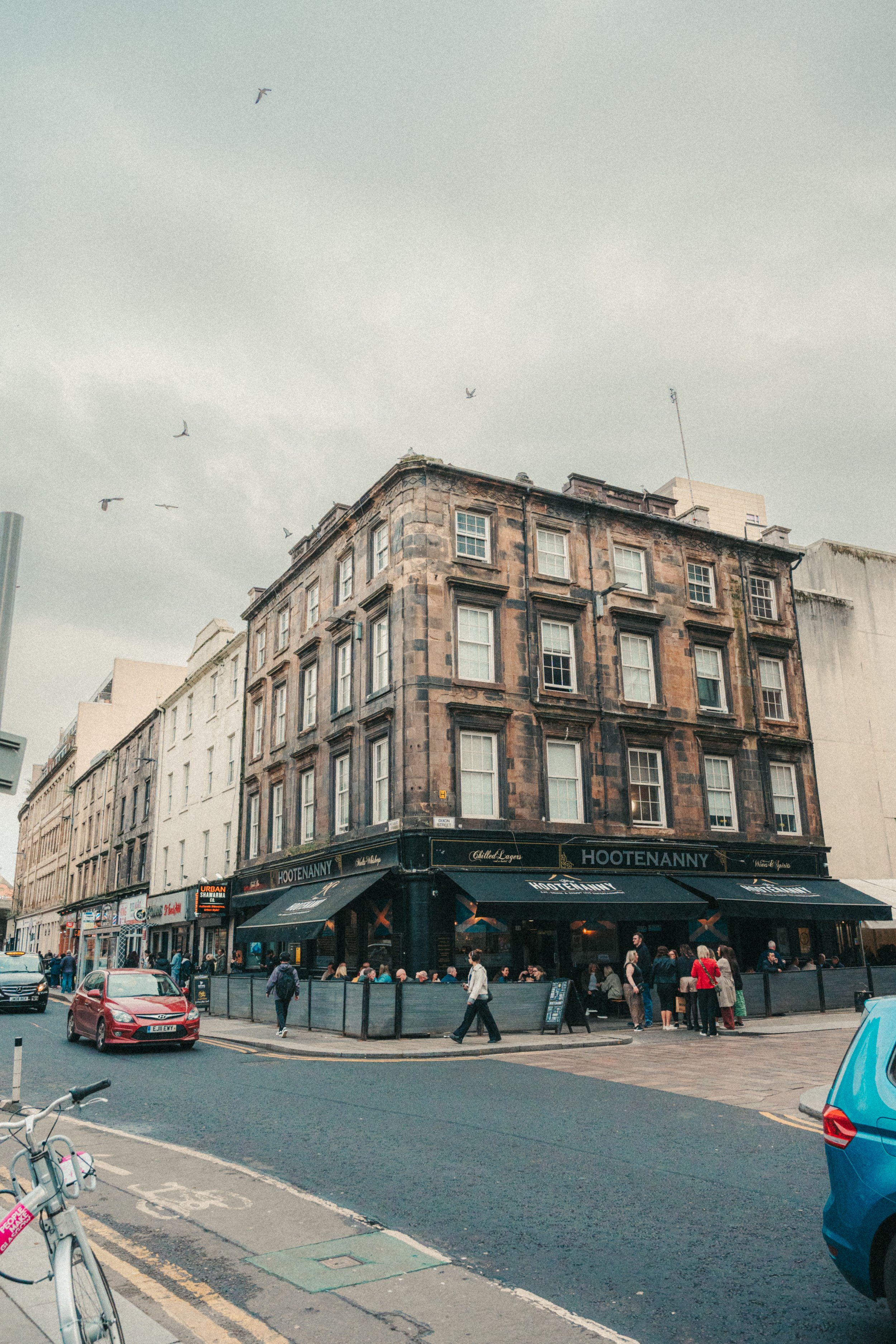 Crowded city street scene with a multi-story brick building at the corner, a busy sidewalk with pedestrians, parked and moving cars, a bicycle in the foreground, and a restaurant called Hootenanny with outdoor seating, and a cloudy sky above.