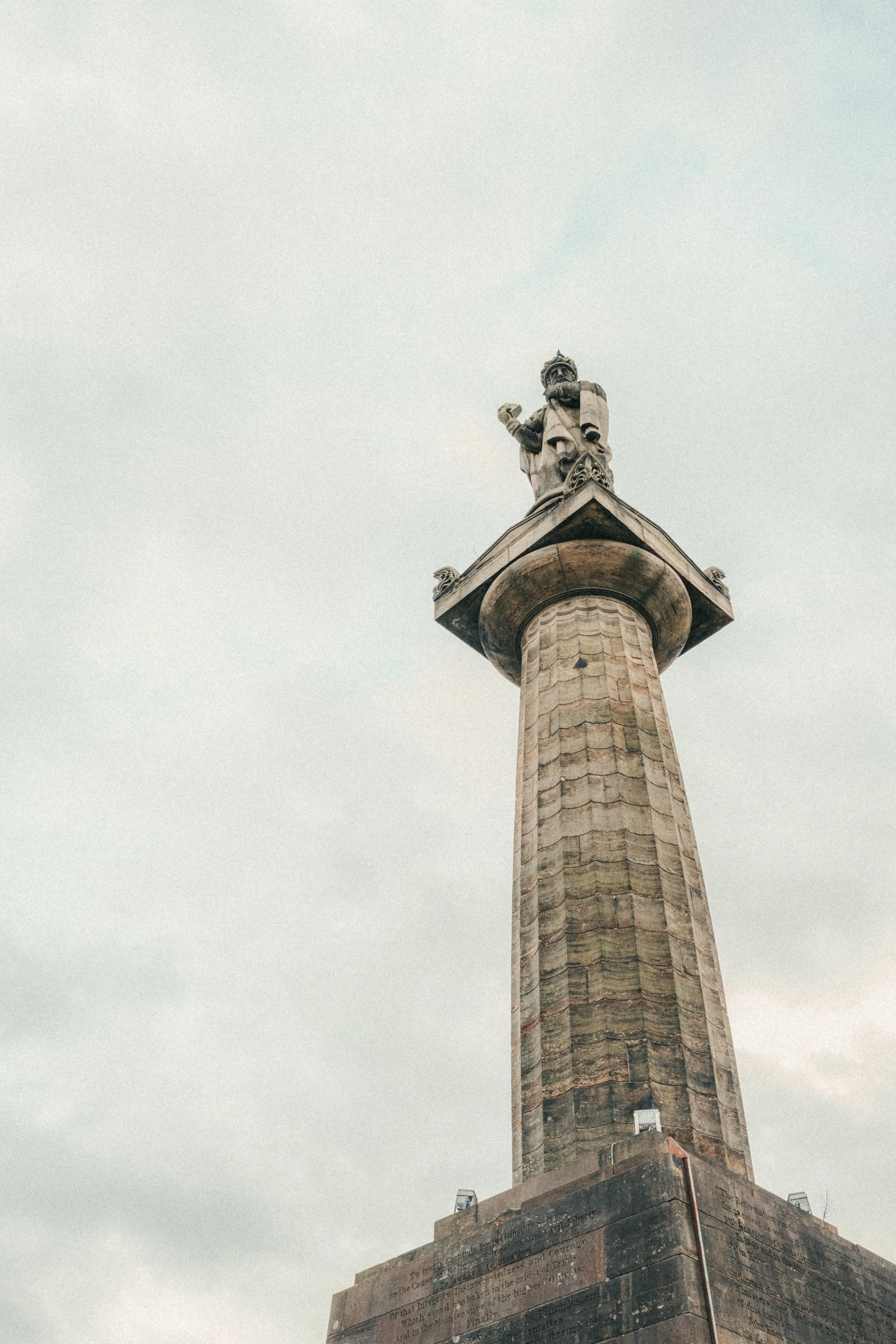A tall stone monument with a statue of a figure holding a scroll and wearing a crown at the top. The monument has inscriptions on its base and is set against a cloudy sky.