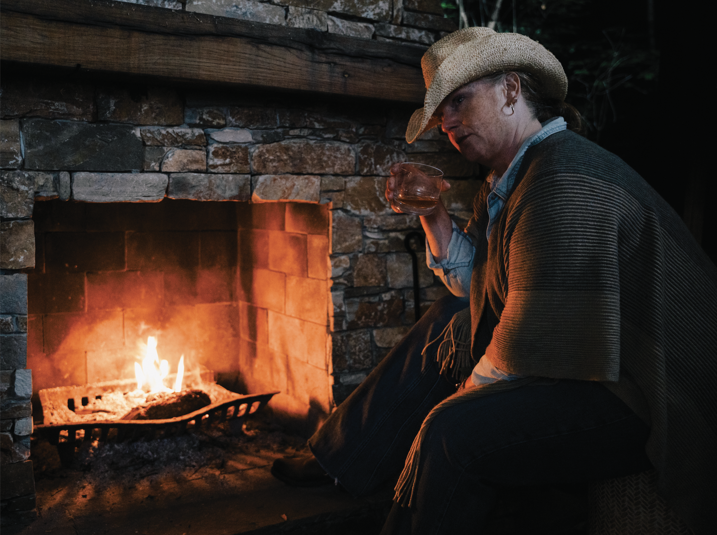 Woman sitting by a lit fireplace, wearing a beige hat, holding a glass of what appears to be wine, and looking down, in a cozy indoor setting.