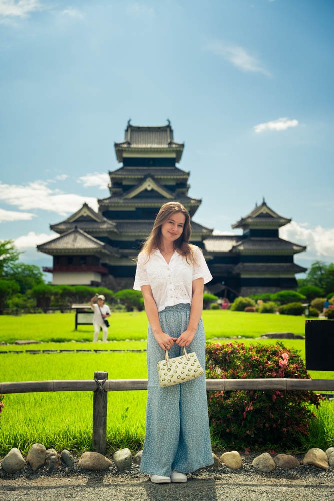A woman standing in front of a traditional Asian pagoda, holding a handbag, with a green lawn and a person taking a photo in the background.