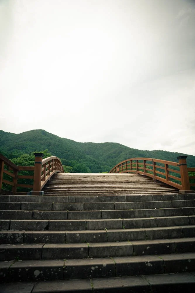 Stone steps leading up to a wooden bridge with green hills and mountains in the background.