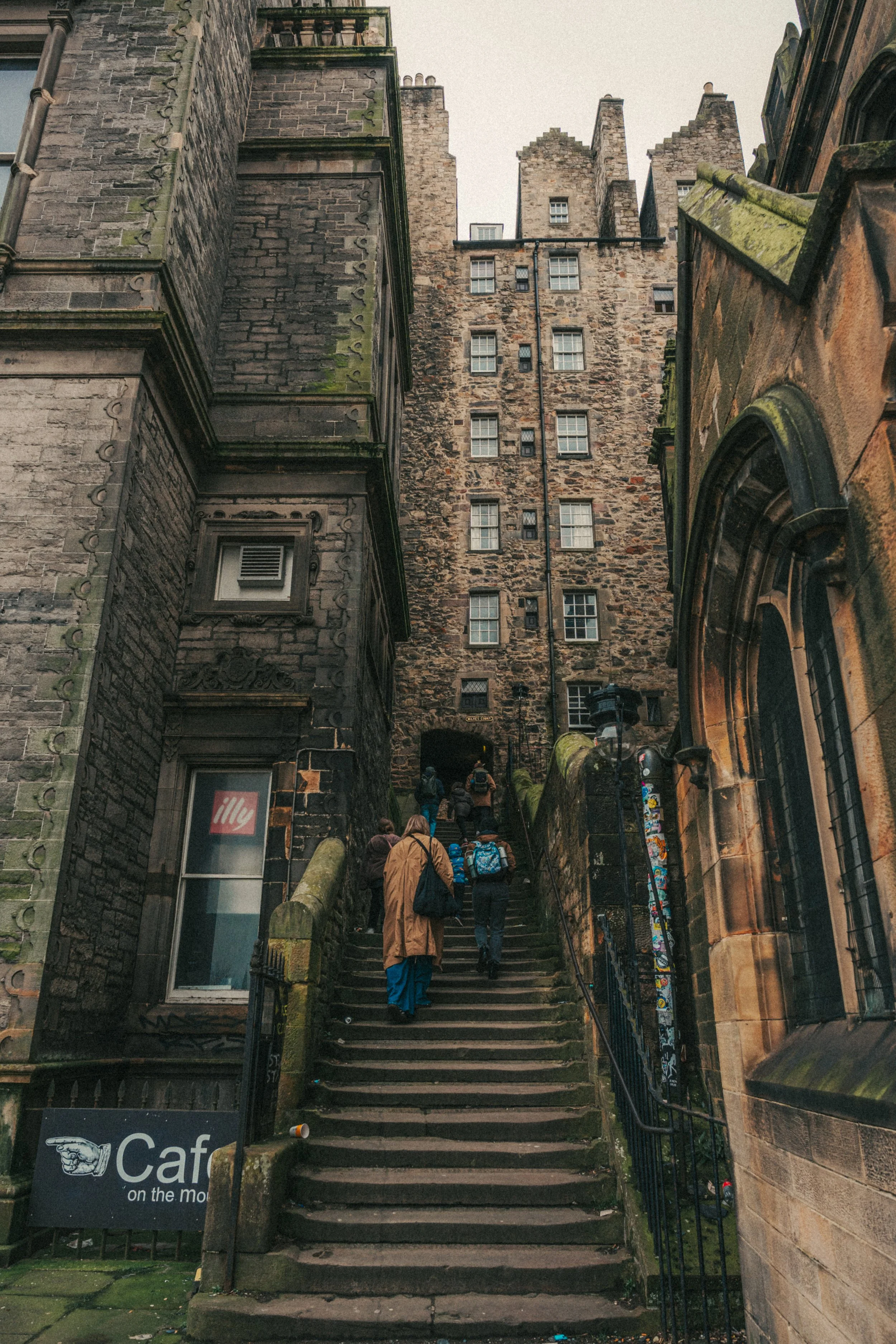 A group of people walking up a set of old, moss-covered stone stairs between historic stone buildings in an urban setting. The photo is taken from the bottom of the stairs looking upward.
