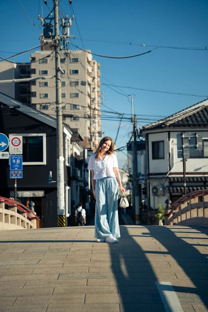 A woman with long hair, wearing a white shirt and light blue wide-leg pants, holding a small handbag, standing on a bridge in an urban area with buildings, power lines, and a clear blue sky in the background.