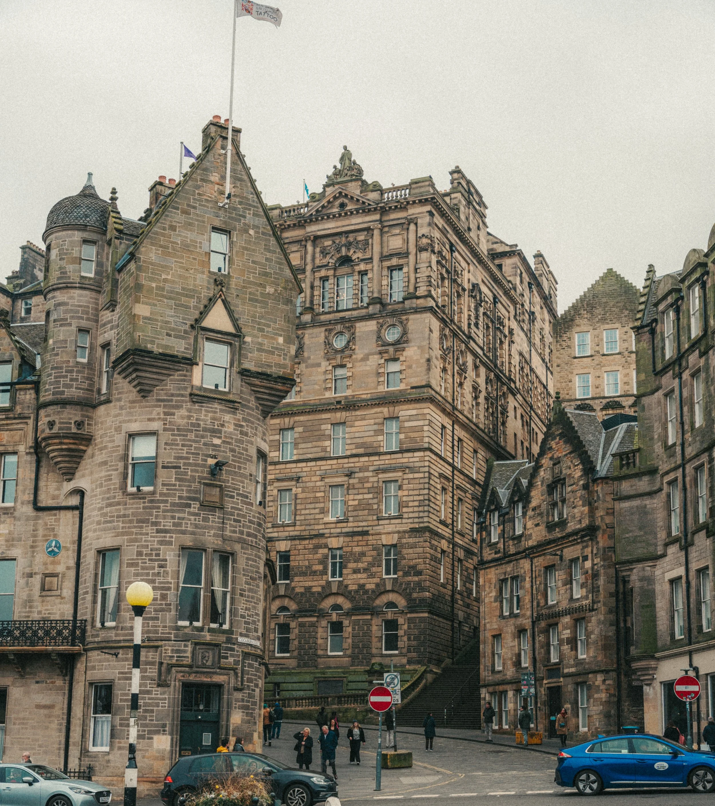 Historic stone building with turrets and flags, outdoor area with people walking, cars parked, signs, and a staircase leading up to another building.