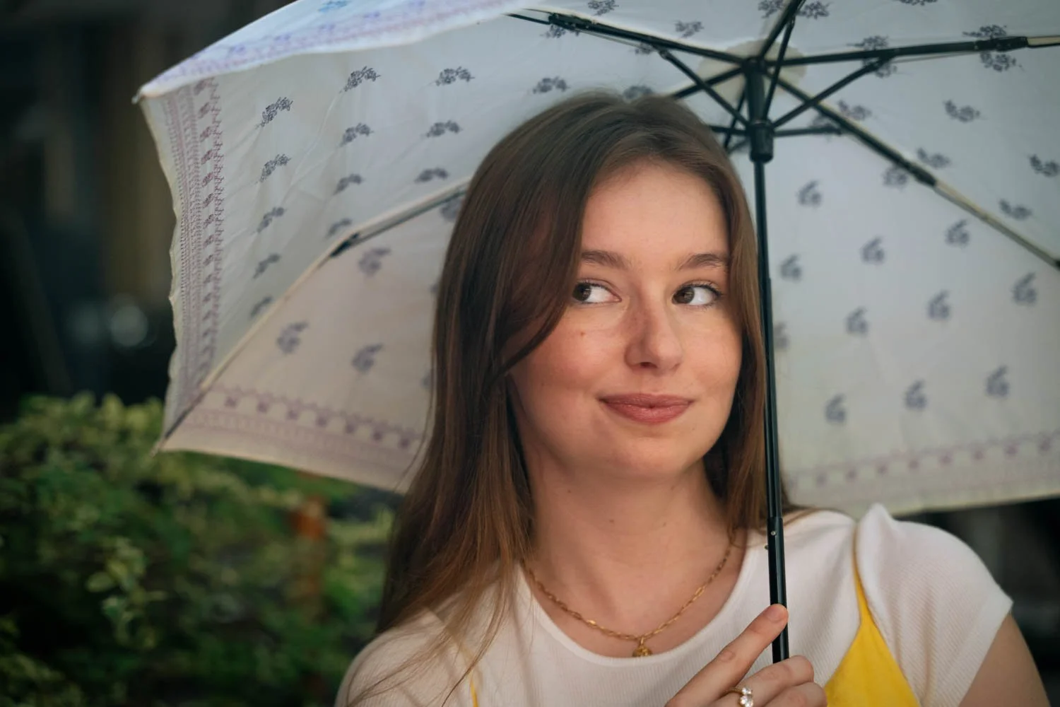 A young woman with brown hair holding a white umbrella with a floral pattern on a cloudy day. She is wearing a white shirt with yellow accents and a gold necklace, smiling softly and looking to her left.