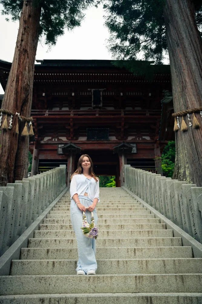 Woman standing on stone steps in front of a traditional Japanese temple, holding a floral bouquet.