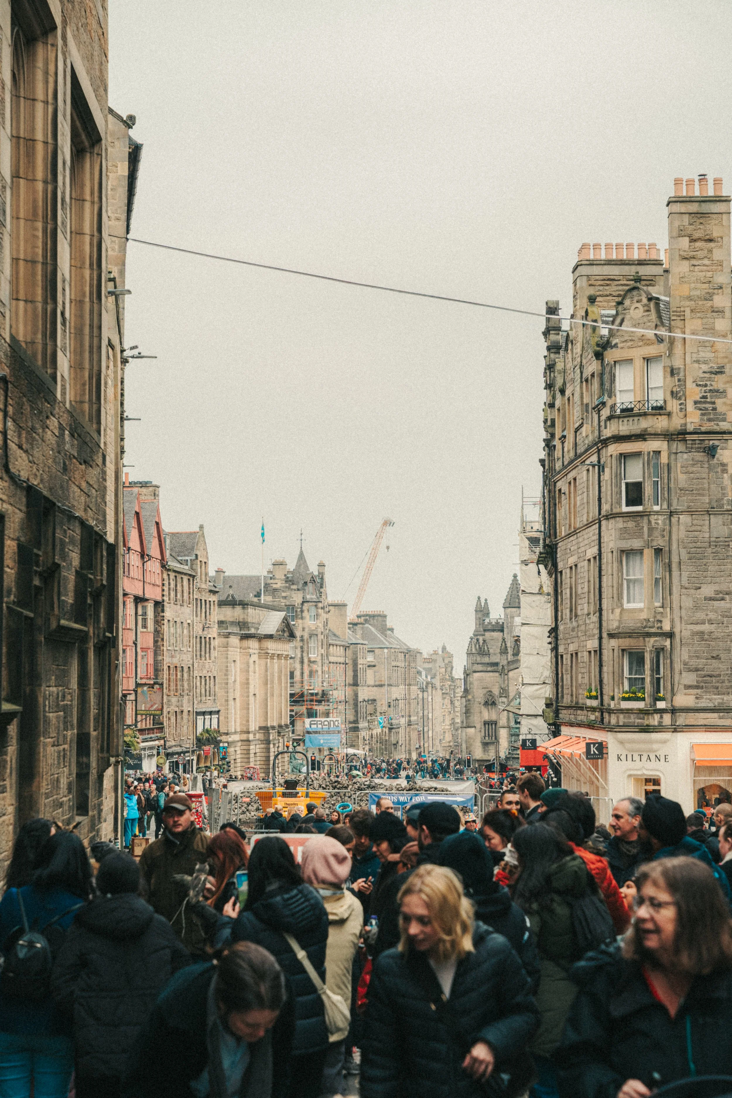A crowded urban street scene with many people walking along the sidewalk. The street is lined with historic stone buildings, some with signs and storefronts, including a store called 'KILTANE'. Overhead wires are visible above the street, and in the 