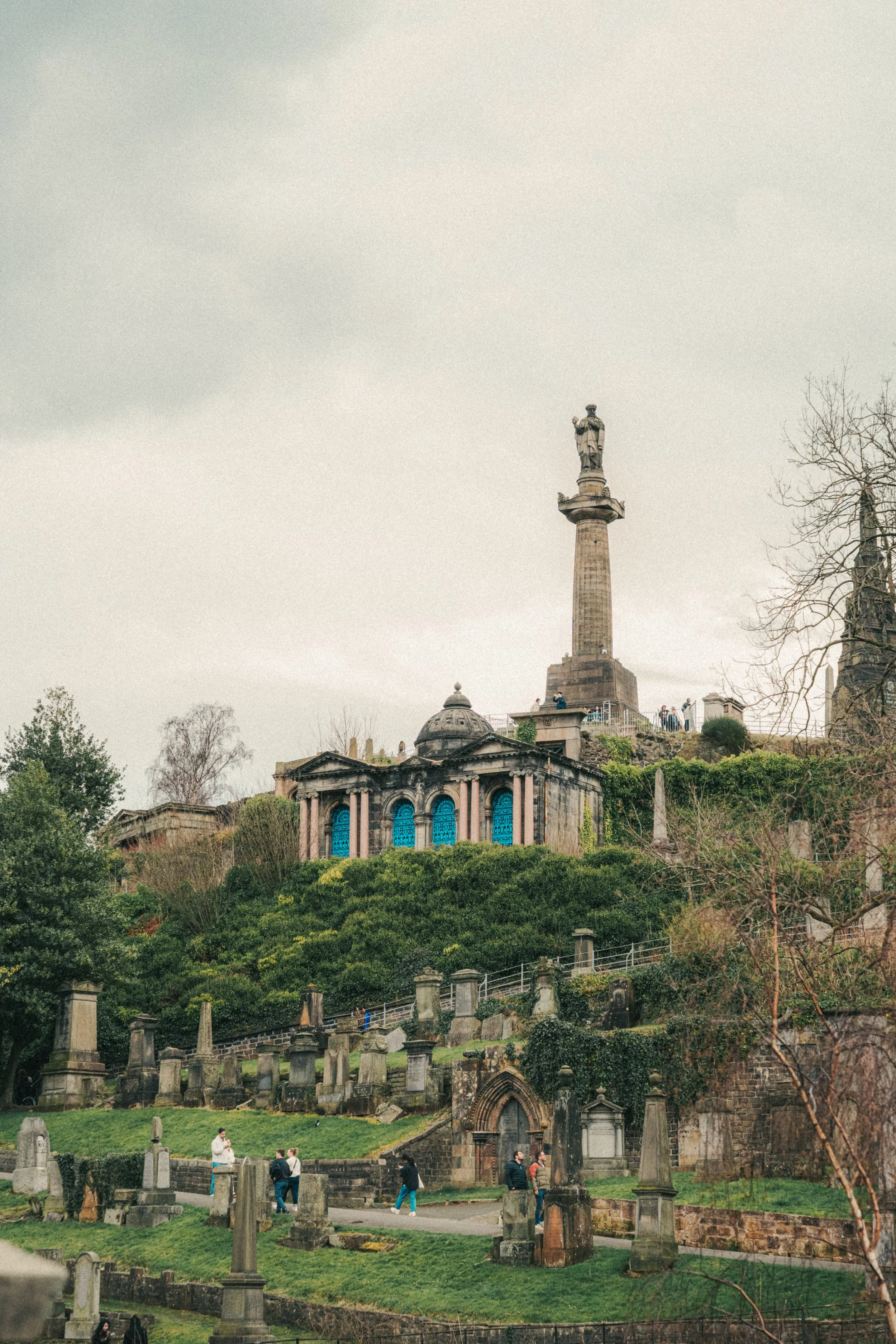 A historic cemetery with numerous tombstones and mausoleums on a grassy slope. In the background, a large monument with a statue of a man on top is situated on a hill, with trees around. Several visitors are walking and exploring the area.