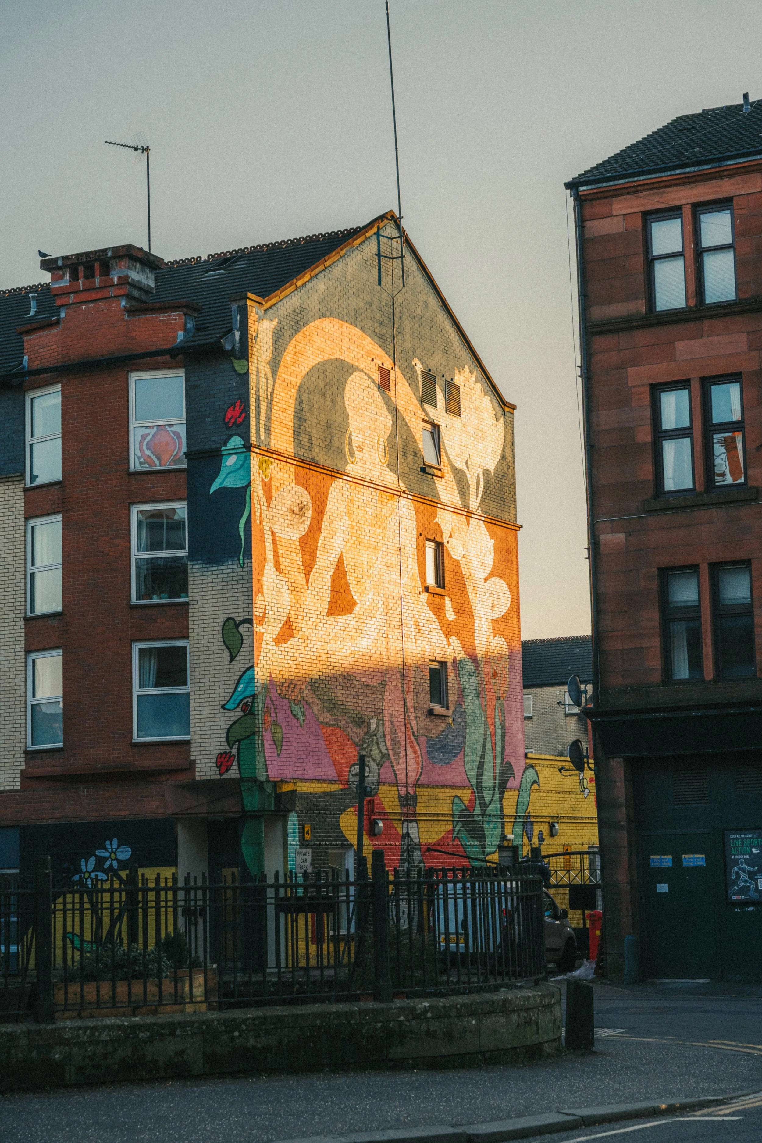 Colorful mural painting of a woman with long hair on the side of a multi-story brick building, with other buildings, a fence, and street in the foreground.