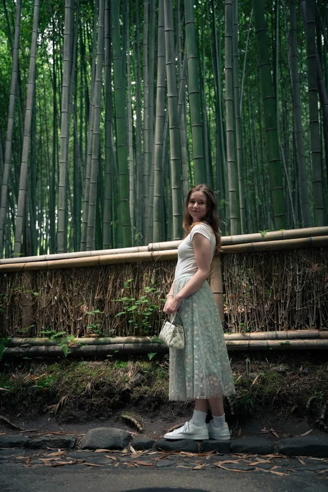 A young woman with light brown hair standing on a path in front of a bamboo forest, holding a small white purse, wearing a white t-shirt, a long lace skirt, and white sneakers.