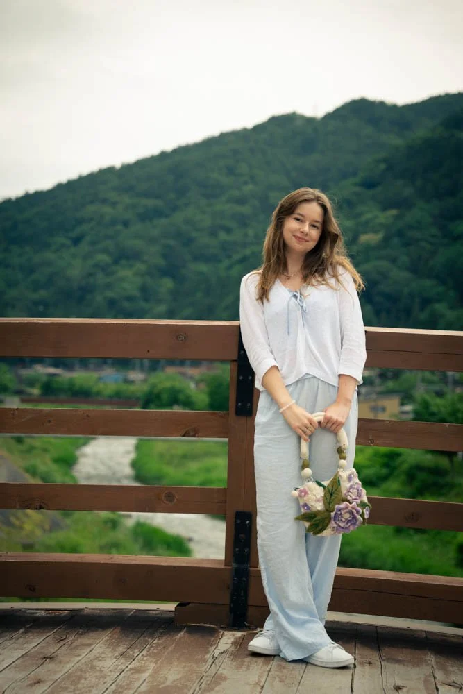 A young woman in white clothing standing on a wooden bridge with a mountain and greenery in the background, holding a floral handbag.