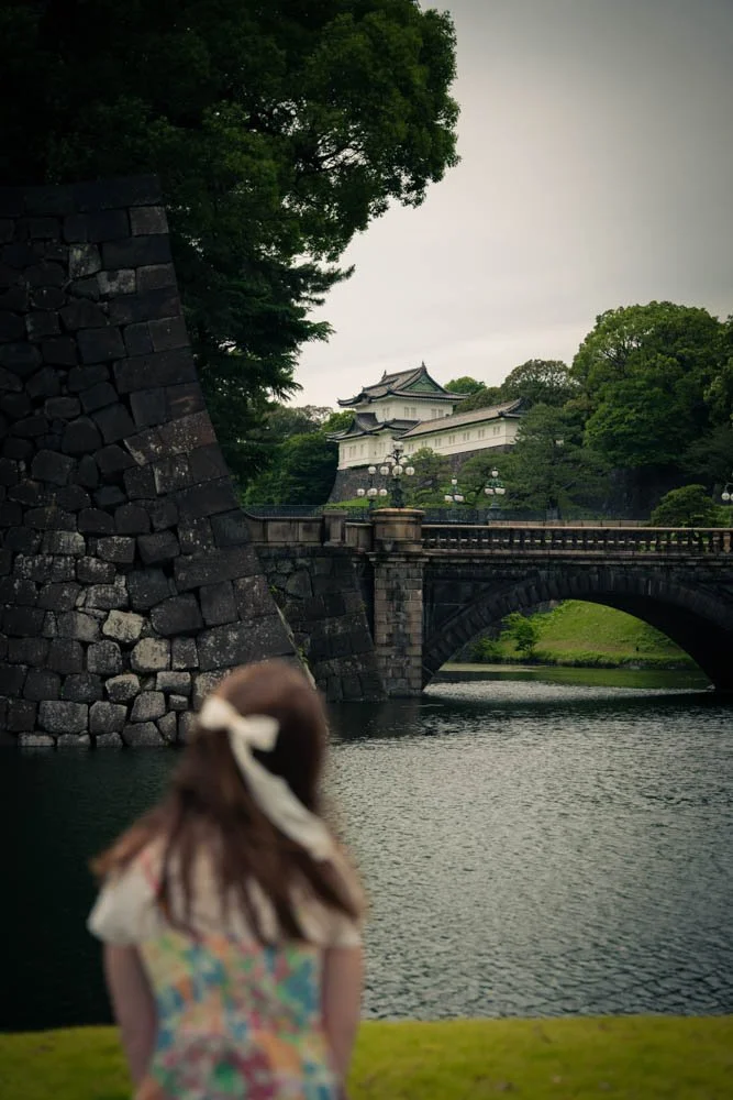 A young girl with a bow in her hair standing on the grassy bank of a river, looking at a traditional Japanese castle in the distance, with a stone bridge and lush green trees.