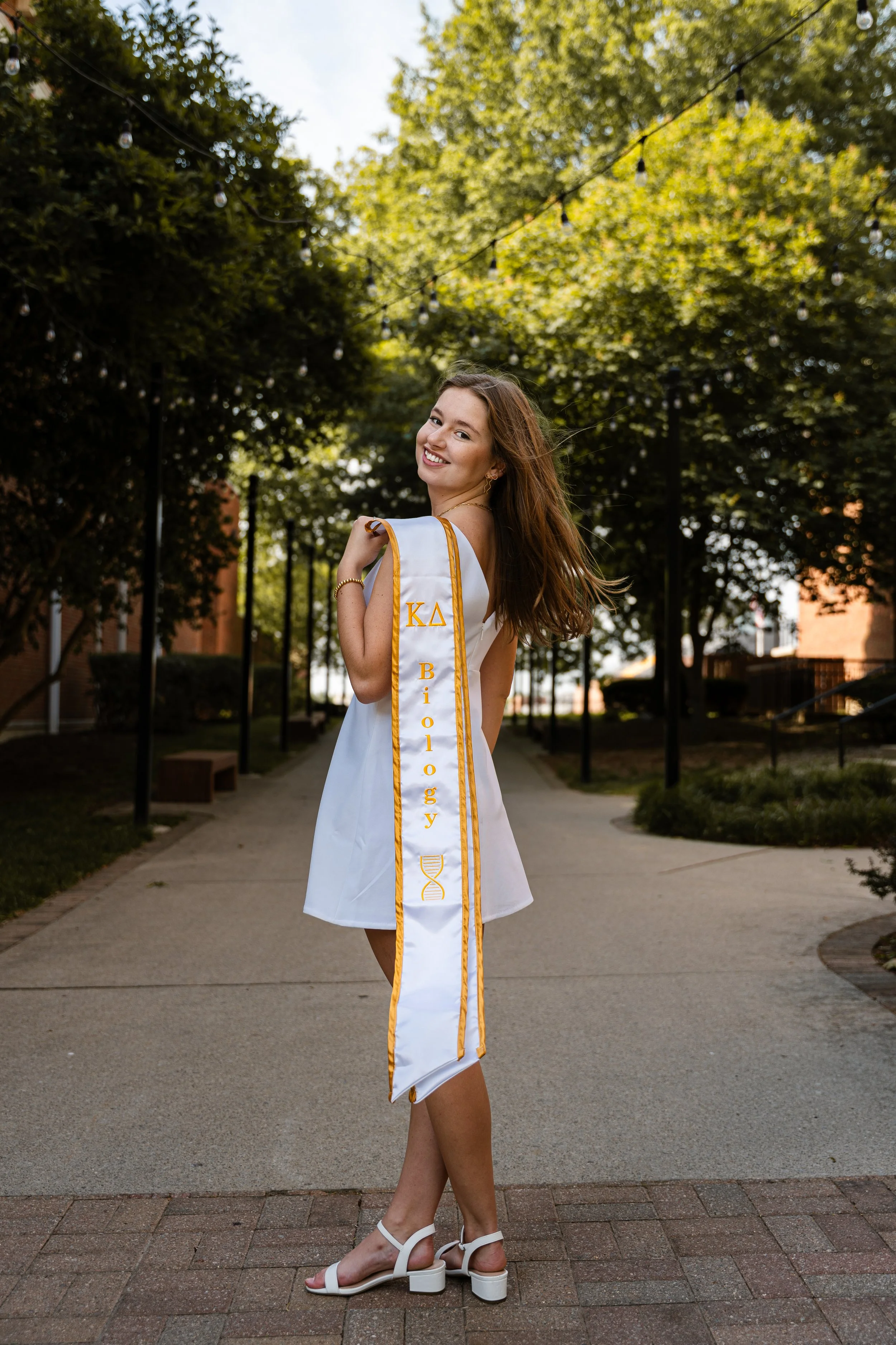 A young woman in a white dress and high-heeled sandals stands outdoors on a paved path, smiling and turning her head over her shoulder. She is wearing a white graduation stole with gold trim that reads 'Kappa Biology' and has a small Greek crest. The