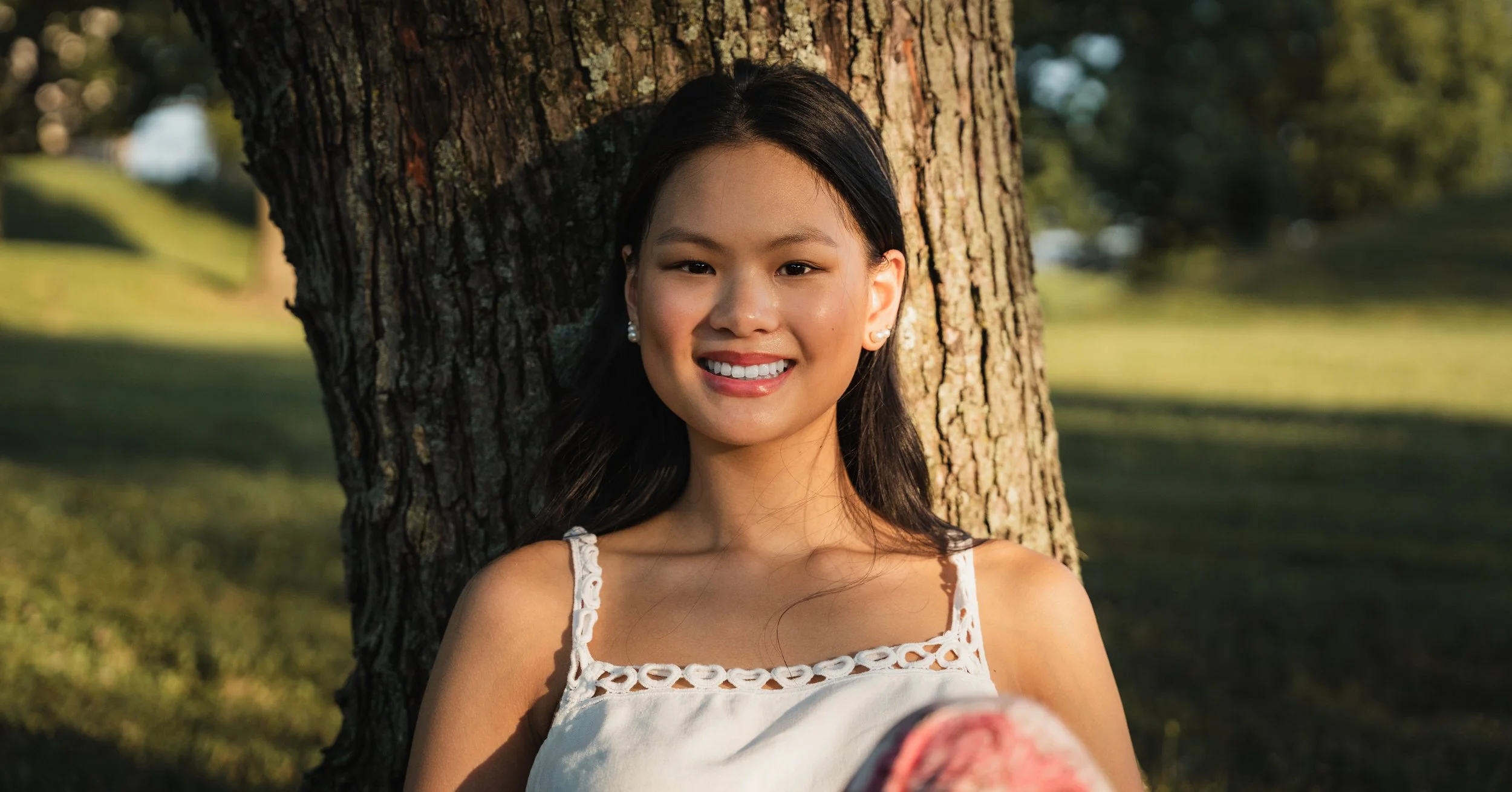 A young woman with long black hair and earrings, smiling, standing against a tree in a park during sunlight.