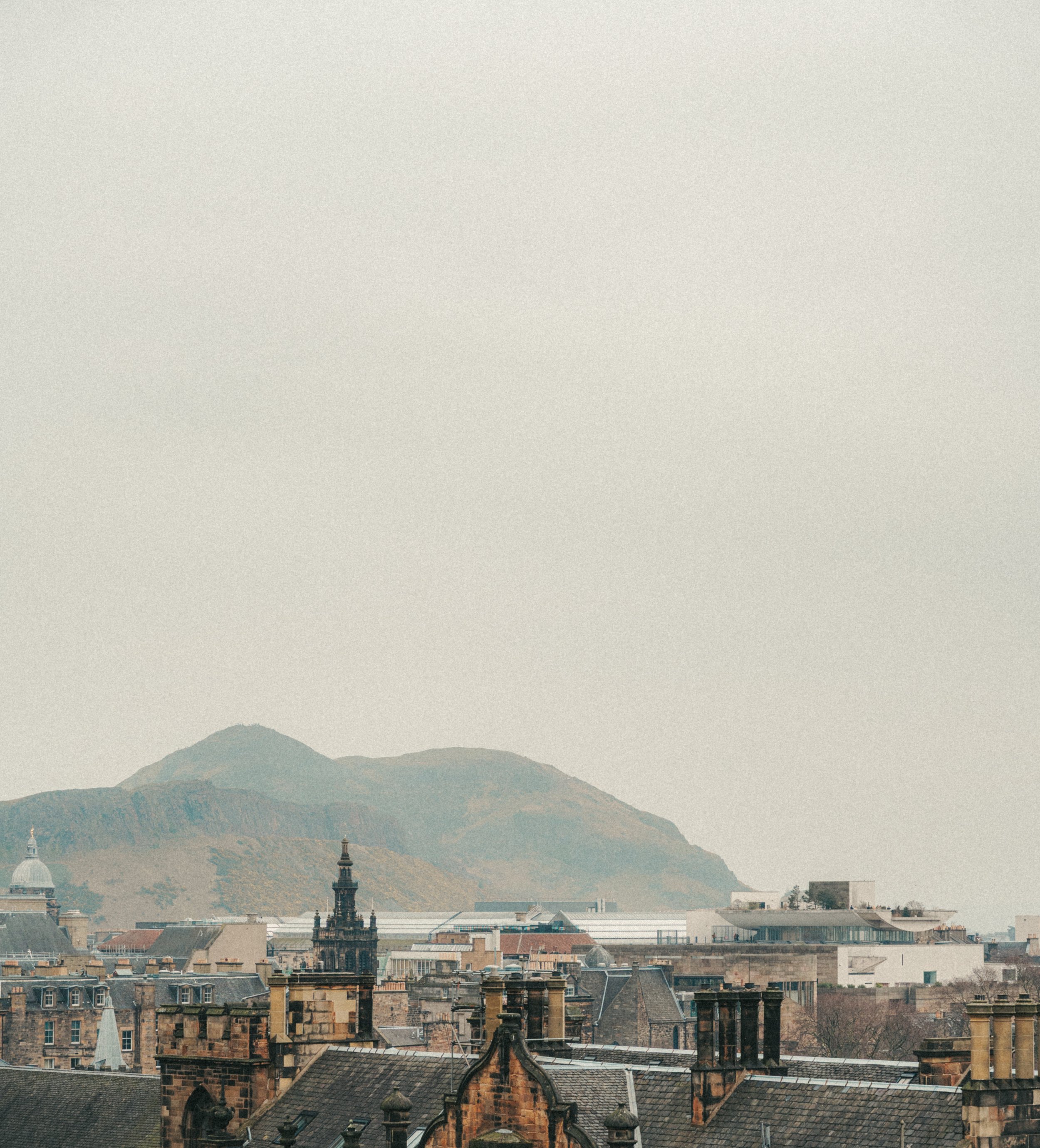 Cityscape with historic buildings and rooftops in the foreground, mountains in the background under a hazy sky.