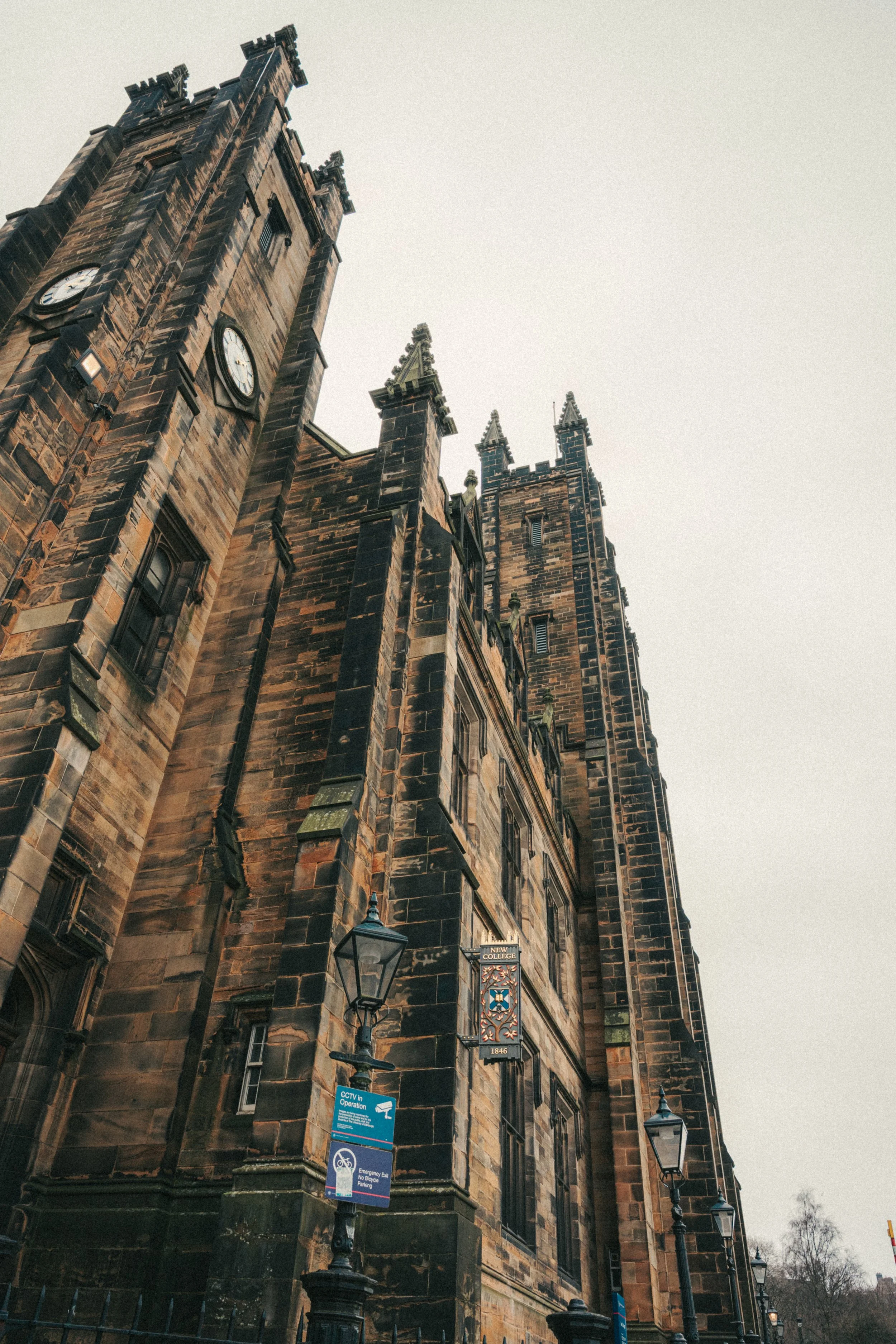 A tall, historic clock tower made of dark brown stone, with ornate gothic architecture, multiple spires, and a clock face showing the time as approximately 1:20, standing against a cloudy sky.