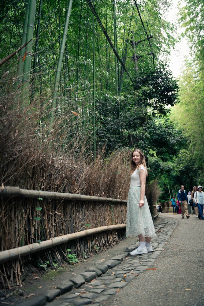 A young woman in a white dress and sneakers standing on a cobblestone path in a lush green park, with a bamboo fence and dense foliage in the background, and some people walking in the distance.