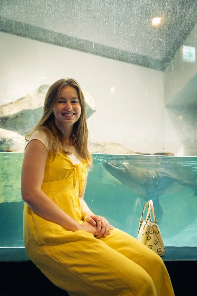 Woman in a yellow dress sitting in front of an aquarium tank with marine life inside at an aquarium.
