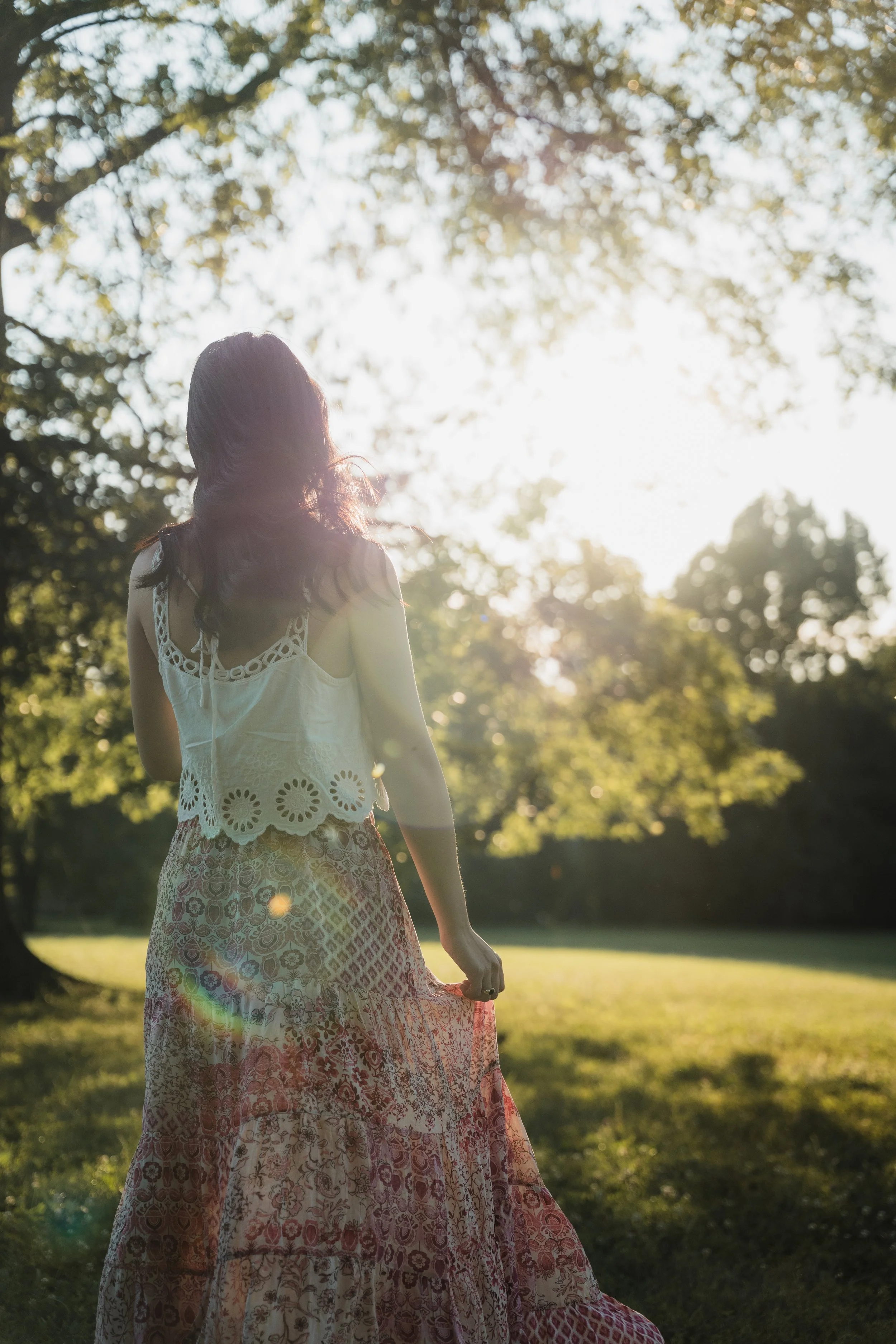 A woman with dark hair standing outdoors in a park, wearing a white sleeveless top with lace details and a long patterned skirt, with sunlight filtering through trees in the background.