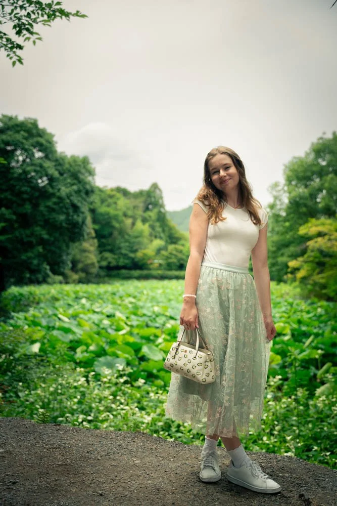 Young woman standing on dirt path beside lush green pond with trees in background, wearing white t-shirt, lace skirt, sneakers, and holding a white purse.