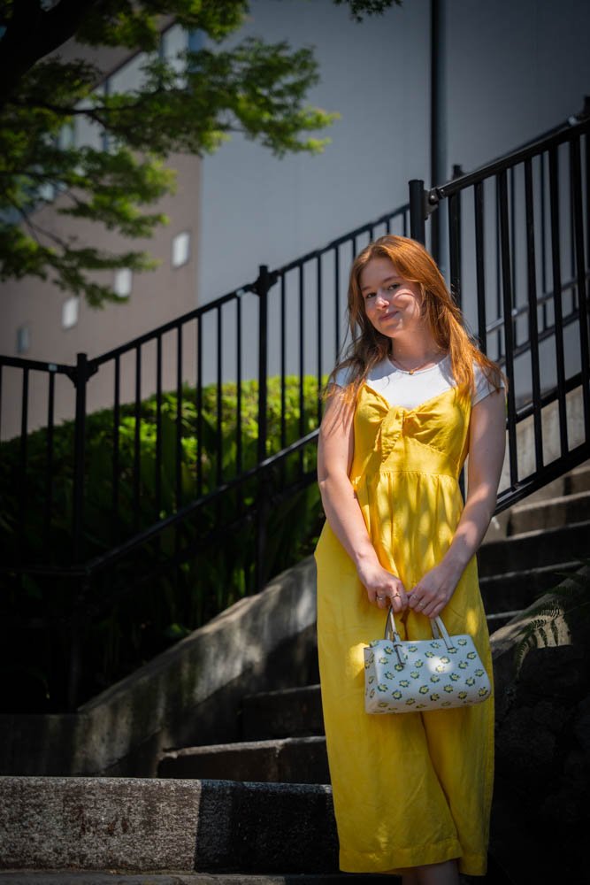 Red-haired woman in a yellow dress holding a patterned handbag standing on stairs outdoors with a black railing and green foliage in the background.